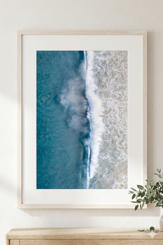 Framed artwork of a beach scene with blue water and white waves on a light wooden surface.