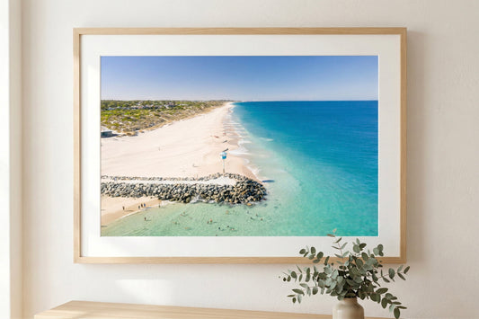 Framed beach scene print on a wall with a plant below