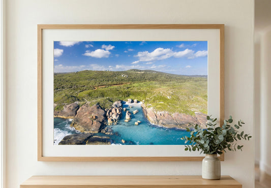 Framed landscape print of a coastal scene with greenery and water, displayed on a wall.
