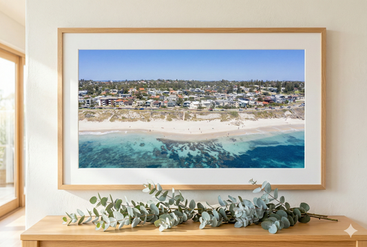 Framed beachscape print on a wall with eucalyptus leaves on a wooden surface below.