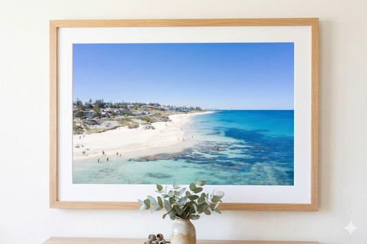 Framed beachscape print on a wall with a vase of flowers below.