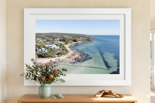 Framed artwork of a coastal scene with a vase of flowers and driftwood on a wooden surface.