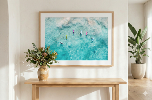 Framed artwork of people in the water on a wall above a wooden table with plants.