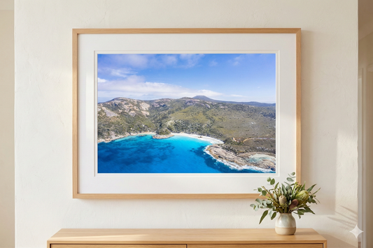 Framed photograph of a coastal landscape with blue water and green hills on a white wall.