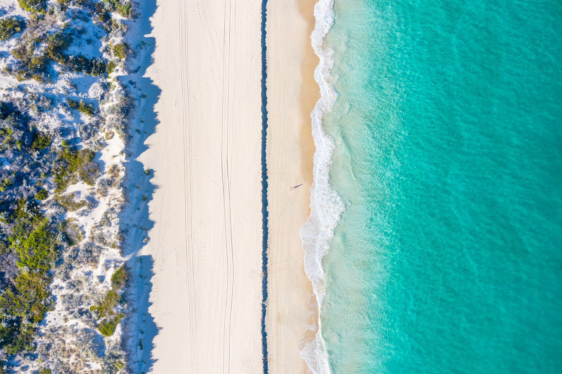 Top-down view of a sandy beach with clear blue water and greenery on one side.