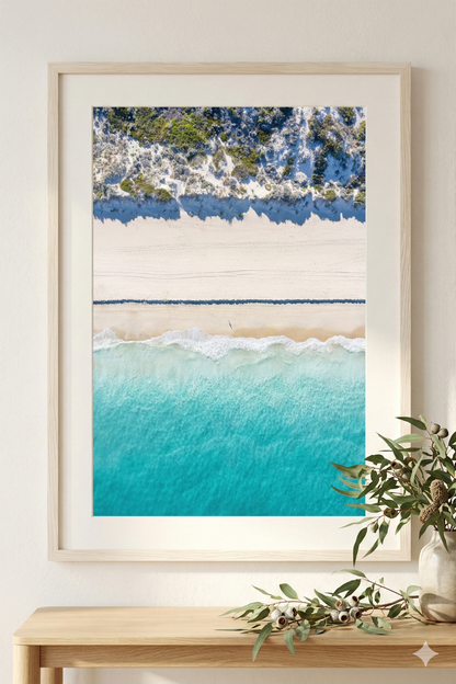 Framed beach scene print on a wall with a plant below