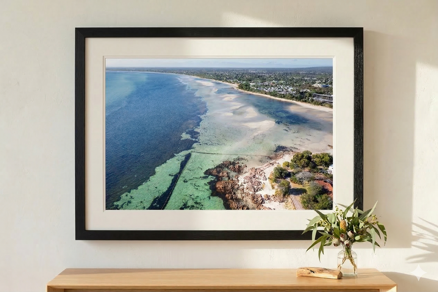 Framed photograph of a coastal scene on a wall above a wooden shelf with a plant.