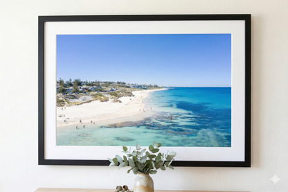 Framed photograph of a beach scene on a wall with a vase and plant in the foreground.