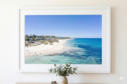 Framed beachscape print on a white wall with a vase and plant below.