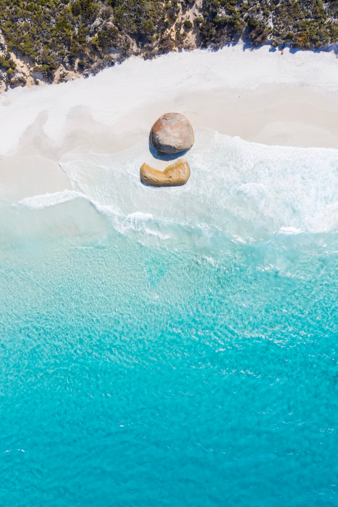 Two stones balanced on top of each other in clear blue water with a sandy beach and greenery in the background.