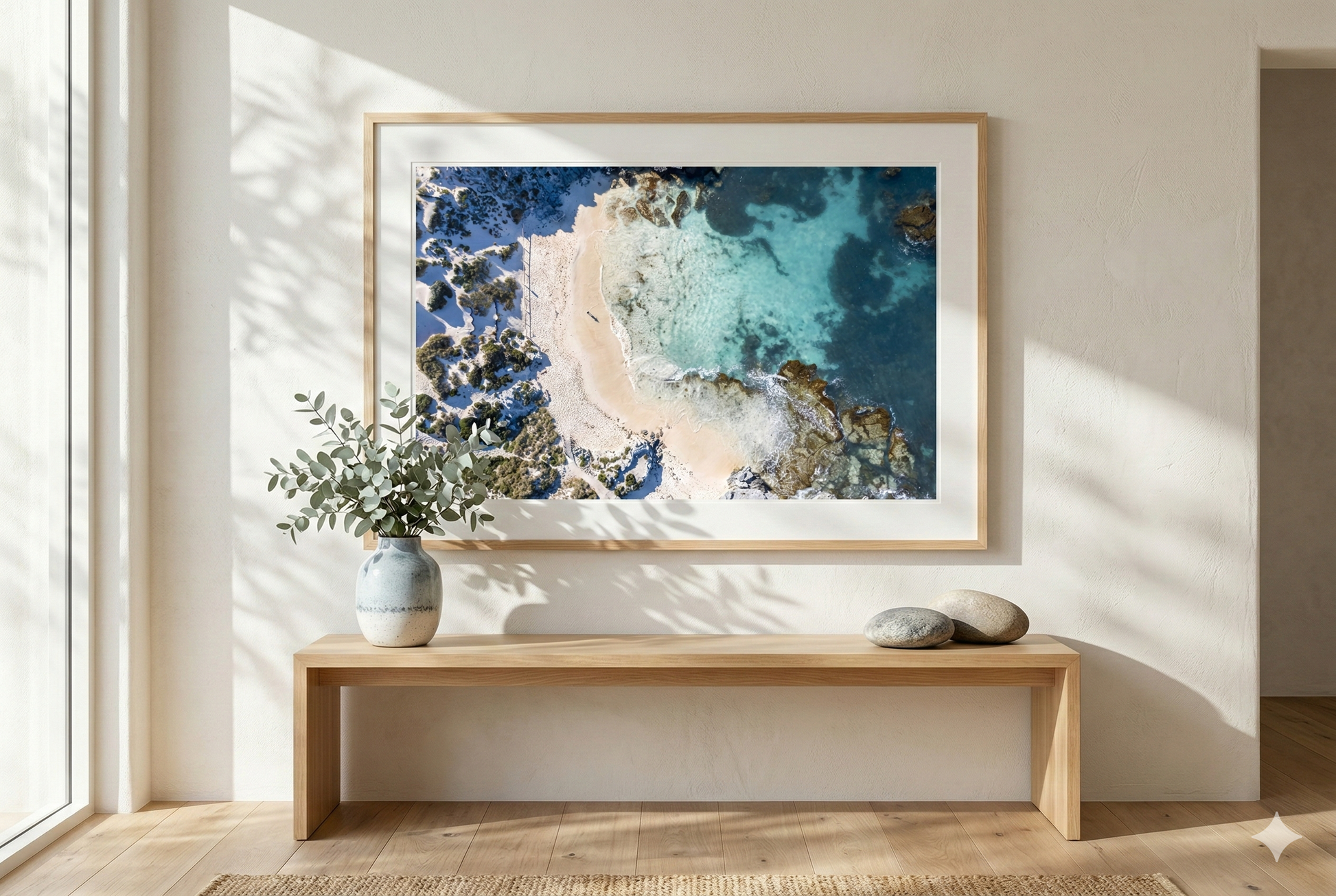 Framed artwork of a coastal scene on a wall in a bright room with a wooden bench and plant.