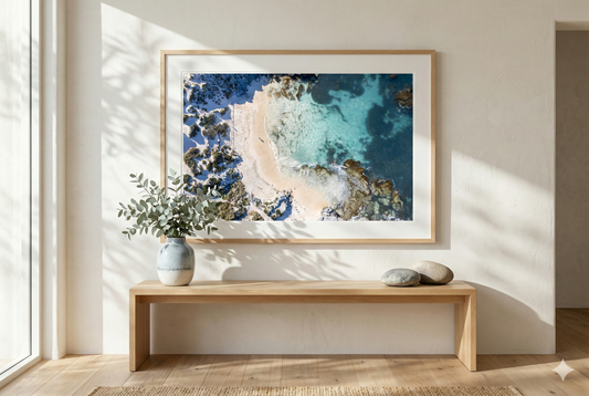 Framed artwork of a coastal scene on a wall in a bright room with a wooden bench and plant.