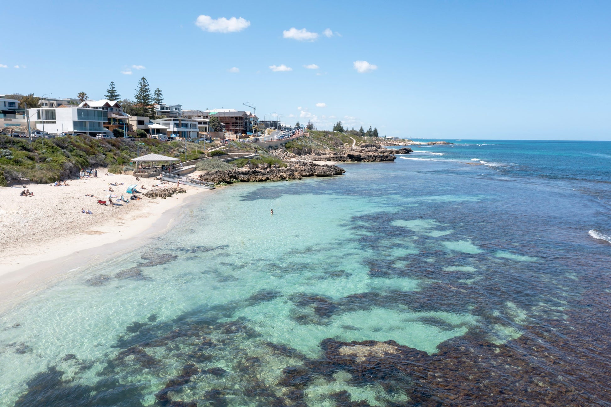 Coastal scene with clear blue water, sandy beach, and houses on a hillside.
