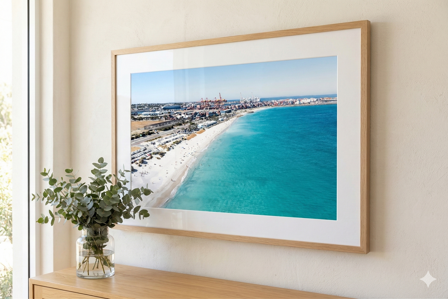 Framed artwork of a beach scene on a wall with a vase of greenery below.