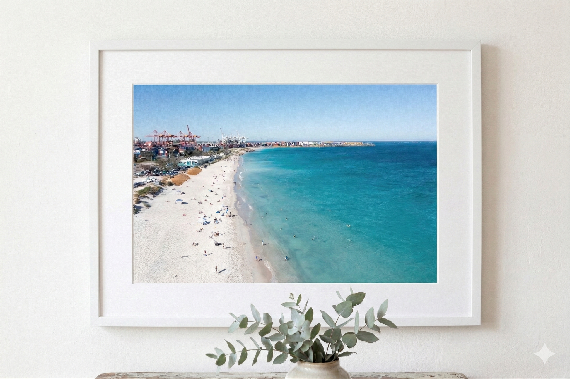 Framed beach photograph on a white wall with a vase of greenery below.