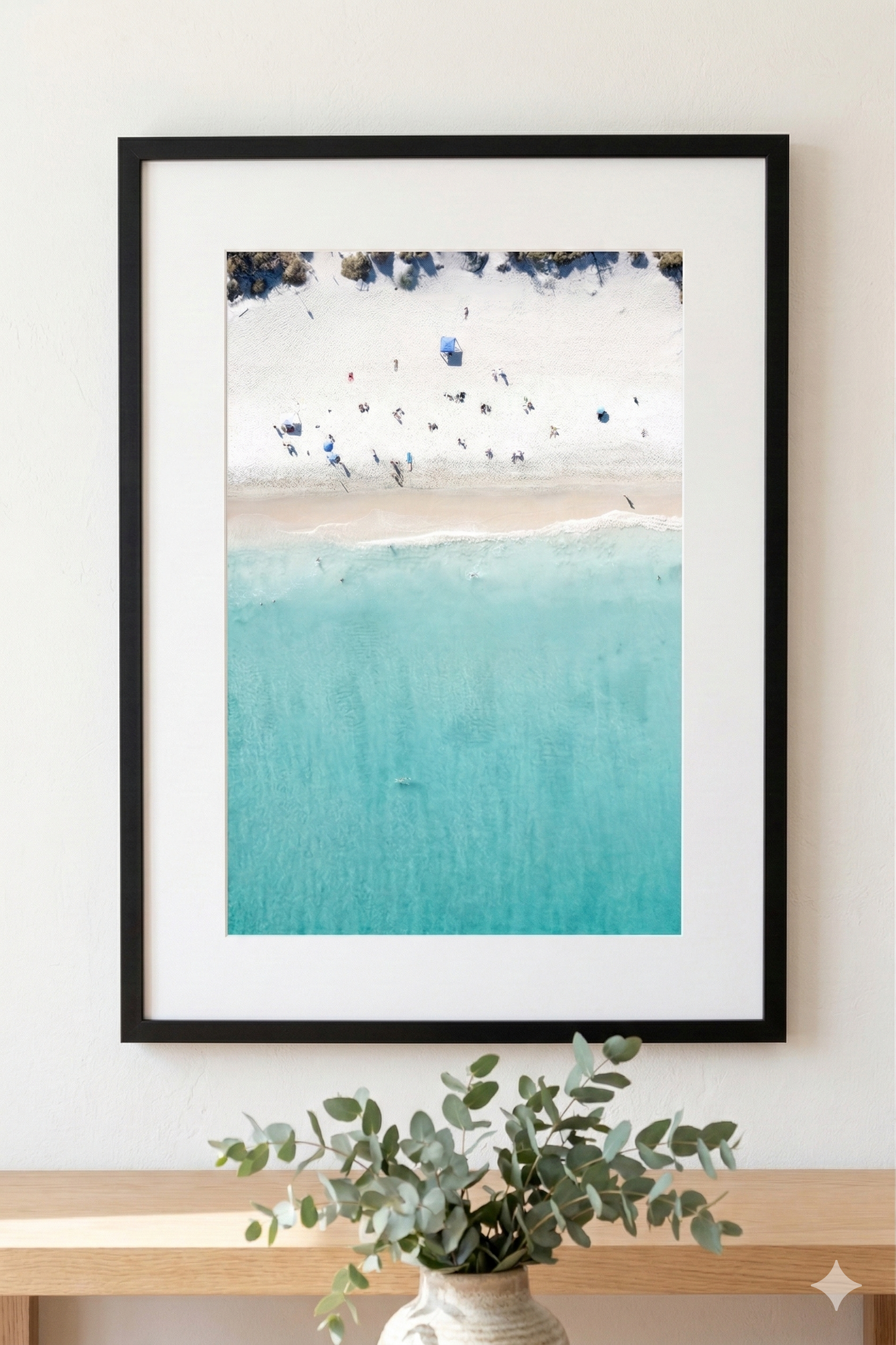 Framed artwork of a beach scene with turquoise water and white sand, displayed on a wall above a wooden shelf with a plant.