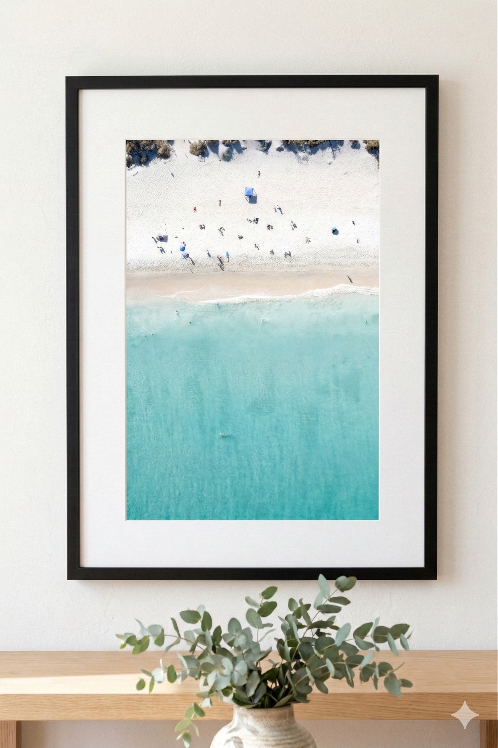 Framed artwork of a beach scene with turquoise water and white sand, displayed on a wall above a wooden shelf with a plant.