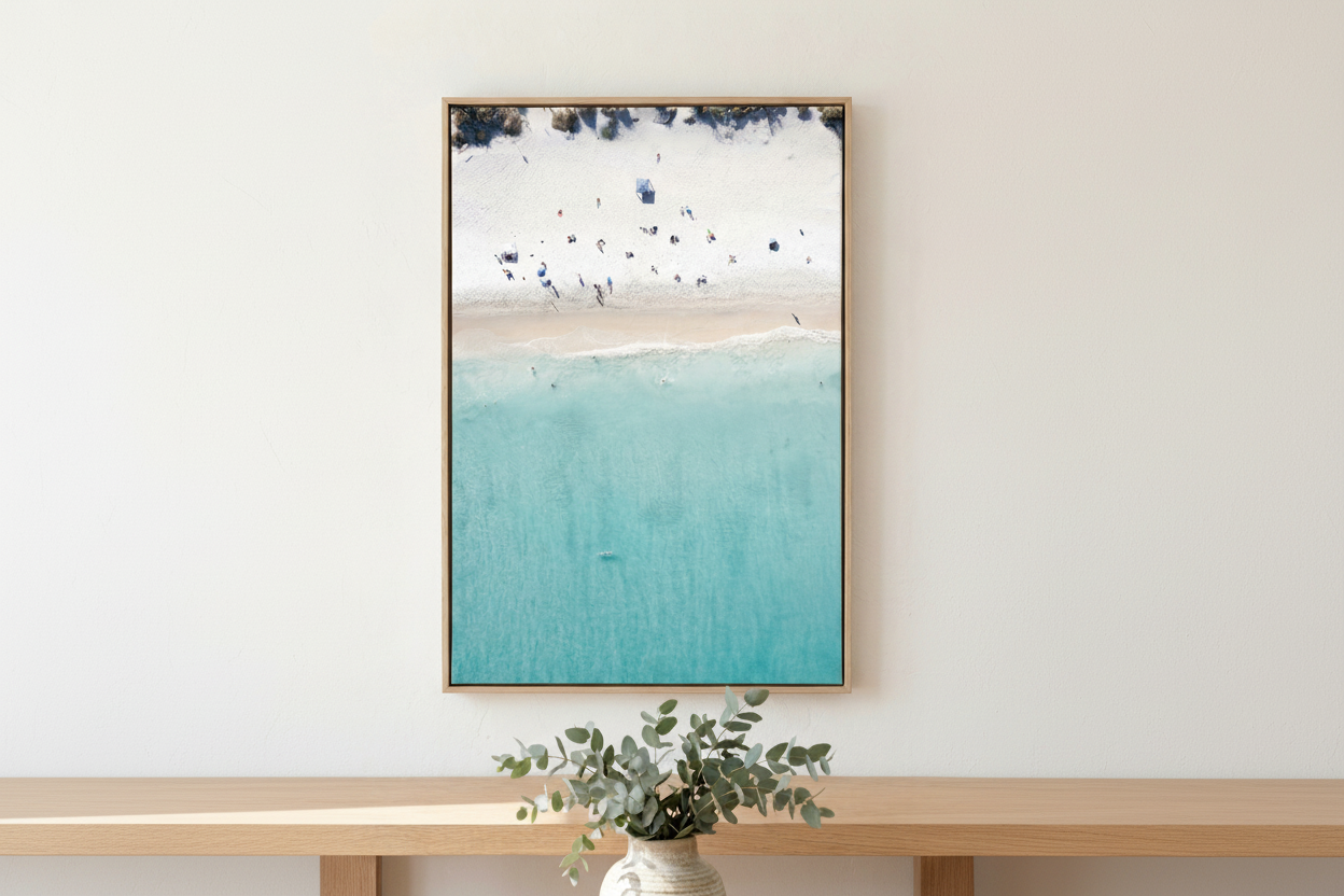 Framed artwork of a beach scene with blue water and white sky, displayed on a wall above a wooden shelf with a plant.