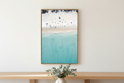 Framed artwork of a beach scene with blue water and white sky, displayed on a wall above a wooden shelf with a plant.