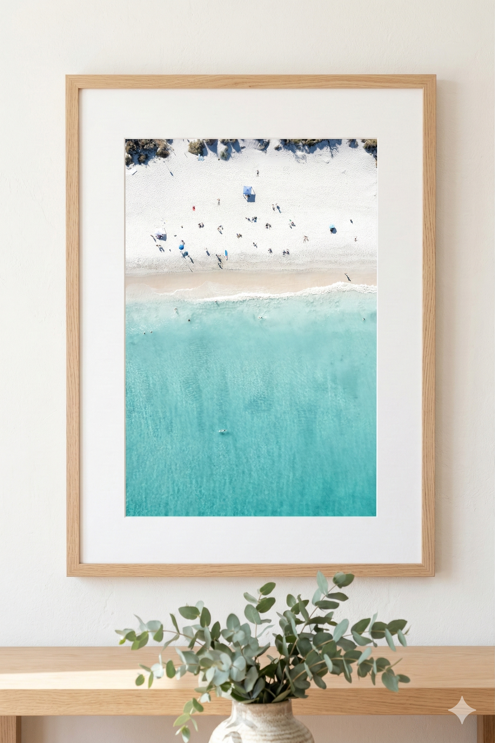 Framed artwork of a beach scene with blue water and white sand, displayed on a wall.