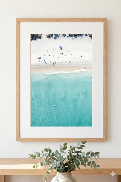 Framed artwork of a beach scene with blue water and white sand, displayed on a wall.