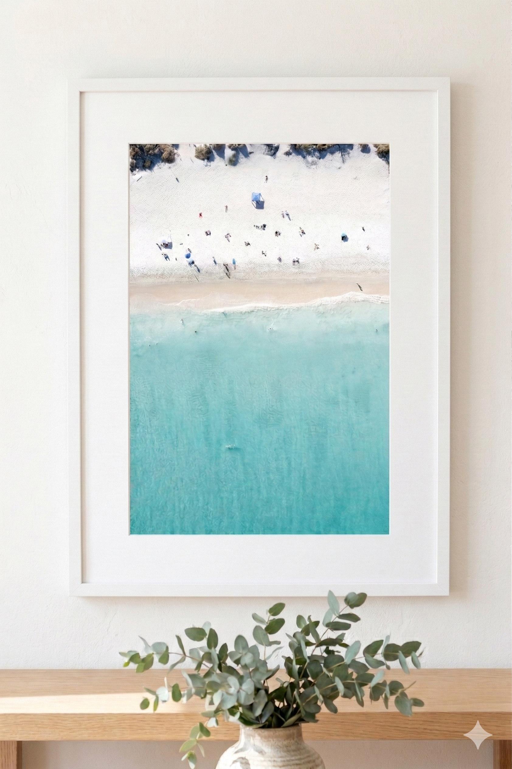 Framed beach scene print on a wall with a vase of eucalyptus below.