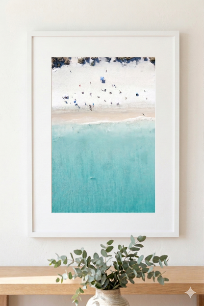 Framed beach scene print on a wall with a vase of eucalyptus below.