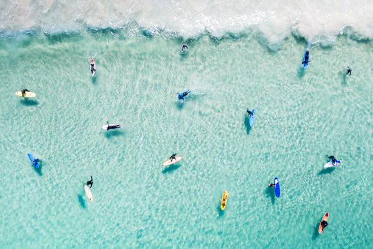 A vibrant fine art print featuring a lively beach scene with multiple people surfing and swimming in the ocean.