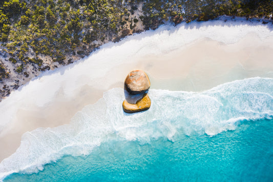 A fine art print featuring a coastal scene with a large rock on the beach and a clear blue water background.